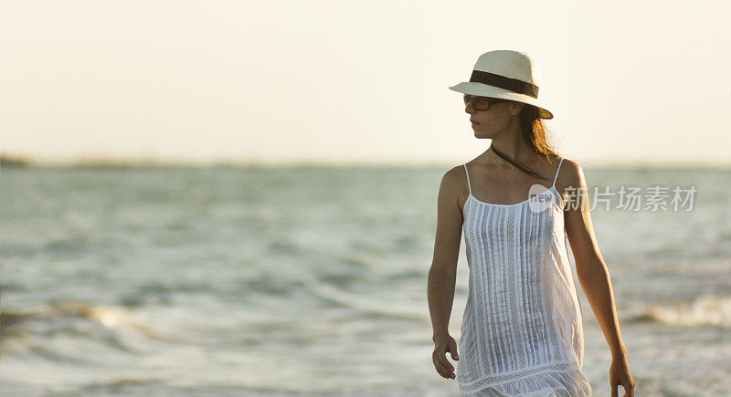 美丽宁静的女人漫步在海滩上，穿着白色连衣裙，草帽和太阳镜在São米格尔dos Milagres，阿拉格阿斯，巴西